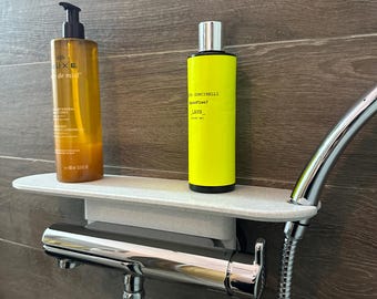 Puede incluir: Una estantería de ducha blanca sostiene dos botellas de gel de ducha. Una es de color ámbar con una bomba negra, la otra es de color amarillo brillante con detalles plateados. La estantería está montada en una pared alicatada, con un cabezal de ducha cromado.