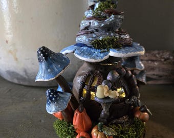 May include: A detailed fairy house sculpture with a spiralling tower, blue mushroom details, and miniature orange pumpkins. The house has a glowing window and rests on green moss, creating a charming miniature scene.