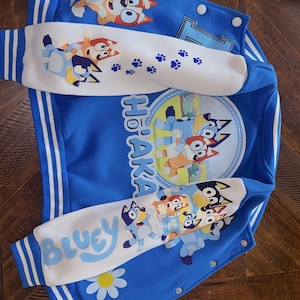 Puede incluir: Una chaqueta estilo universitario de color azul brillante con los personajes Bluey y Bingo. La chaqueta tiene mangas blancas con huellas de patas azules y el texto Bluey. Un gr&aacute;fico circular muestra a los personajes y el texto Ho'aka impreso.