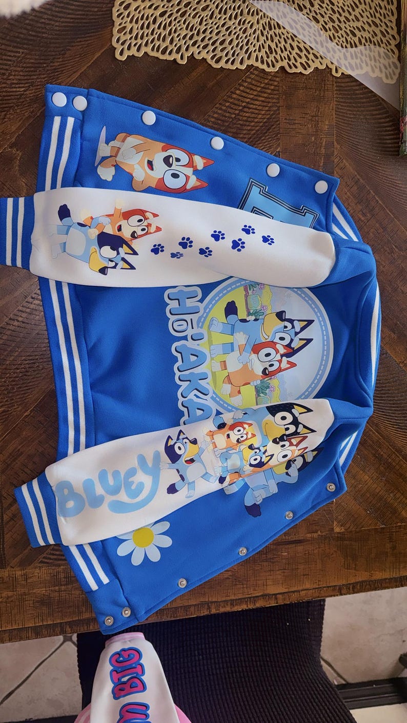 Puede incluir: Una chaqueta estilo universitario de color azul brillante con los personajes Bluey y Bingo. La chaqueta tiene mangas blancas con huellas de patas azules y el texto Bluey. Un gr&aacute;fico circular muestra a los personajes y el texto Ho'aka impreso.
