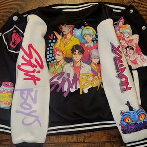 Puede incluir: Una chaqueta universitaria negra y blanca con un estampado gr&aacute;fico de cinco personajes estilo anime en la espalda. Las mangas son blancas con texto rosa y morado que dice Saja Boys y Huntrax, junto a ilustraciones de latas y un gato.