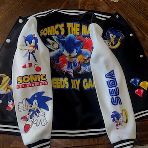 Puede incluir: Una chaqueta estilo universitario en blanco y negro con gr&aacute;ficos de Sonic the Hedgehog. La parte trasera muestra a Sonic con el texto SONIC'S THE NAME. Las mangas presentan personajes de Sonic, el logo de SEGA y anillos dorados.