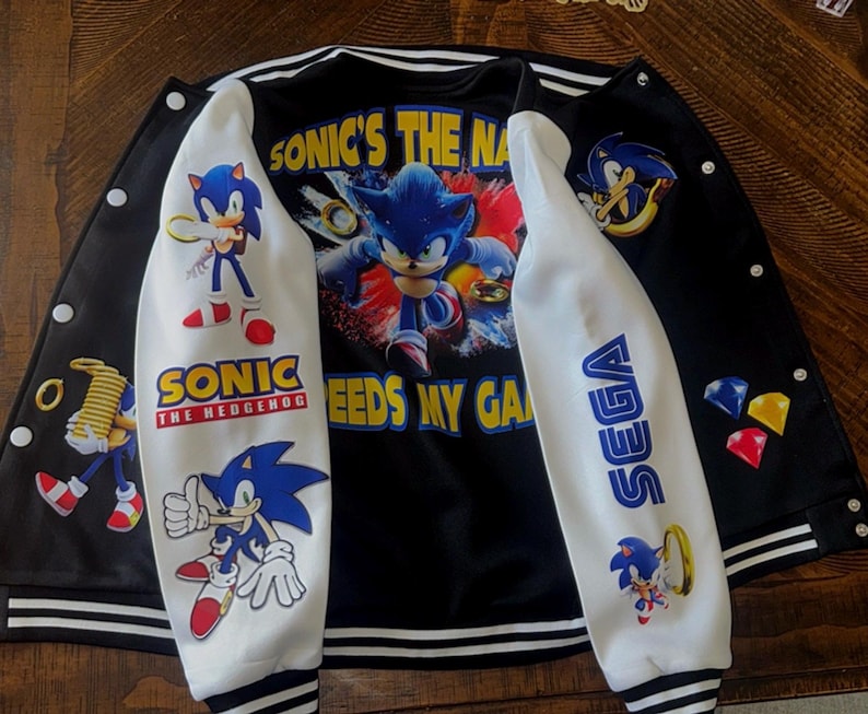 Puede incluir: Una chaqueta estilo universitario en blanco y negro con gr&aacute;ficos de Sonic the Hedgehog. La parte trasera muestra a Sonic con el texto SONIC'S THE NAME. Las mangas presentan personajes de Sonic, el logo de SEGA y anillos dorados.