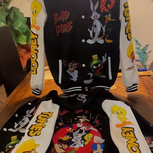 Puede incluir: Una chaqueta universitaria en blanco y negro con personajes de Looney Tunes como Bugs Bunny, el Pato Lucas, Piol&iacute;n, Marvin el Marciano y Speedy Gonzales. La chaqueta tiene mangas blancas con texto Looney Tunes, capucha y botones a presi&oacute;n.
