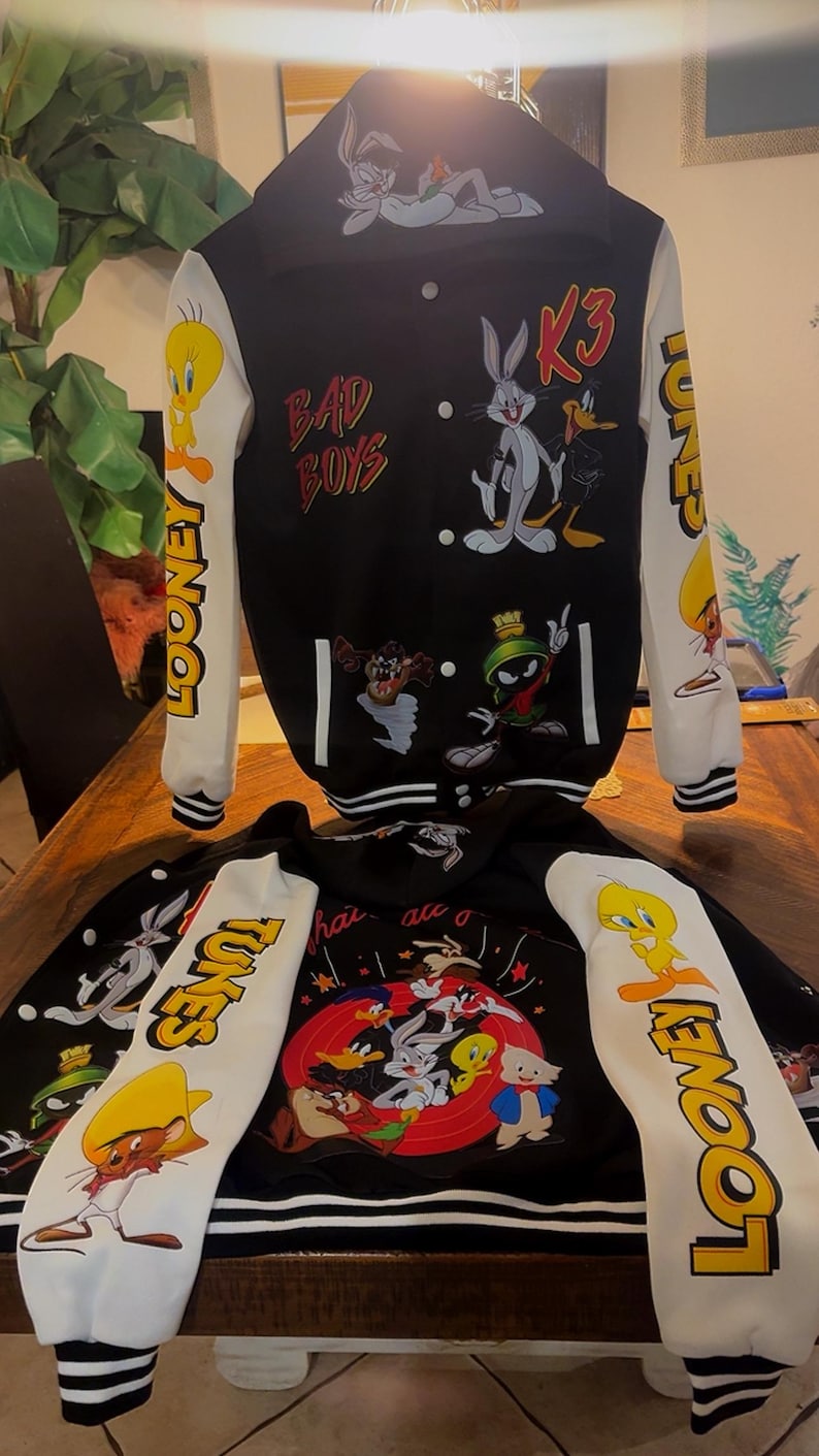 Puede incluir: Una chaqueta universitaria en blanco y negro con personajes de Looney Tunes como Bugs Bunny, el Pato Lucas, Piol&iacute;n, Marvin el Marciano y Speedy Gonzales. La chaqueta tiene mangas blancas con texto Looney Tunes, capucha y botones a presi&oacute;n.