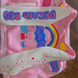 Puede incluir: Una chaqueta estilo universitario rosa con personajes de Peppa Pig, un arco&iacute;ris y un sol amarillo. Las mangas blancas muestran el texto DREAM BIG en letras azules y un patr&oacute;n de estrellas rosas. La chaqueta tiene botones a presi&oacute;n blancos.