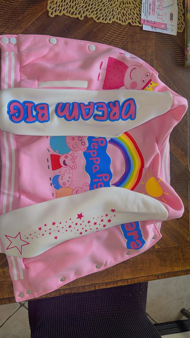 Puede incluir: Una chaqueta estilo universitario rosa con personajes de Peppa Pig, un arco&iacute;ris y un sol amarillo. Las mangas blancas muestran el texto DREAM BIG en letras azules y un patr&oacute;n de estrellas rosas. La chaqueta tiene botones a presi&oacute;n blancos.
