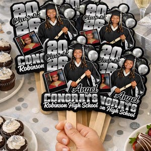 May include: A hand holding graduation fans with the text "Class of 2026" and "Congrats Angel Robinson High School on High School". Each fan features a photo of a graduate in a cap and gown, with black and silver accents.