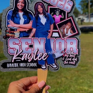 May include: A graduation photo prop featuring photos of a person, the text "Class of 2026", "Senior Kaylee", and "Bayside High School". The prop is silver glitter with a wooden handle.