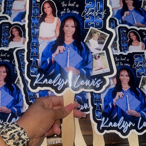May include: A collection of graduation fans featuring a young woman in a blue graduation gown. The fans have the text "Congrats" and "Class of 2025" with the name "Kaelyn Lewis". The fans are held together with wooden sticks.