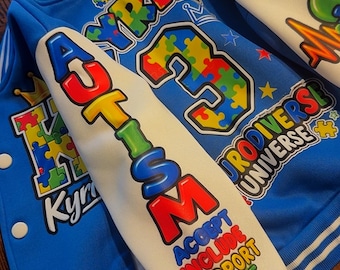 Puede incluir: Una chaqueta universitaria azul con mangas blancas que presentan gráficos de piezas de rompecabezas coloridas, coronas y estrellas. El texto dice AUTISM, KYRIE, 3, NEURODIVERSE UNIVERSE, ACCEPT, INCLUDE, SUPPORT y LOVE. Botones a presión blancos.