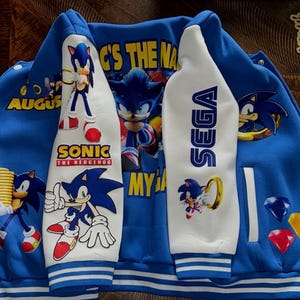 May include: Blue and white Sonic the Hedgehog varsity jacket. The jacket features images of Sonic, the text "SONIC THE HEDGEHOG", and the SEGA logo. The sleeves are white with blue accents. The jacket has a button-up front and a striped hem.