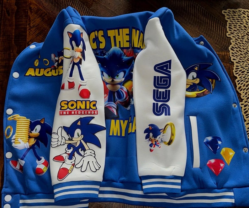 Puede incluir: Una chaqueta universitaria azul y blanca con gr&aacute;ficos de Sonic the Hedgehog. El frente muestra a Sonic con anillos dorados, el texto 'SONIC THE HEDGEHOG', 'SEGA' y 'AUGUST'. La parte posterior muestra a Sonic con el texto 'G'S THE NAME' y 'MY GA'.