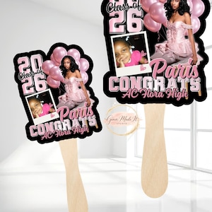 May include: Two graduation fan decorations. Each fan features a photo of a young woman, pink balloons, and the text "Class of 26" and "Paris Congrats AC Flora High" in pink and black. The fans have wooden handles.