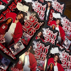 May include: A stack of graduation announcements with a black border. The announcements feature a photo of a graduate in a red graduation gown, the text "Class of 2026", and the graduate's name. A small note says "I did it!"