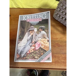以下が含まれることがあります： パム・ドーソン著のヴィンテージ「Knitting Fashion」の本。表紙にはピクニックを楽しむカップルが描かれており、「A Step by Step Guide to Knitting & Crochet」と「115 Patterns to Knit and Crochet for All the Family」と書かれています。