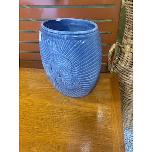 May include: A blue ceramic vase with a seashell design. The vase is cylindrical with a wide opening and features a raised seashell and starfish design on the front. The vase is sitting on a wooden surface.
