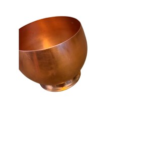 Könnte beinhalten: Eine abgerundete Kupferschale mit glatter, metallischer Oberfläche und einem kreisförmigen Boden. Die Schale hat einen warmen, rotbraunen Farbton und eine polierte Oberfläche, die das Licht reflektiert und ein schlichtes, geschwungenes Design zeigt.