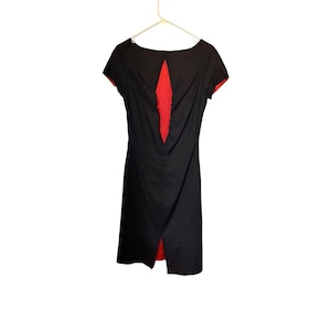 May include: A black knee-length dress with short sleeves and a boat neckline. The back features a vertical diamond-shaped cutout revealing a bright red lining. The dress includes a central zipper closure and a small slit at the bottom hemline.