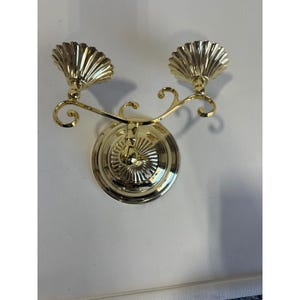 May include: A gold-colored wall sconce with two candle holders. The sconce features a decorative, scalloped design around the candle holders and a circular base. The arms curve gracefully, adding to its ornate appearance. The sconce is mounted on a white wall.