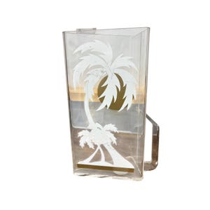May include: A clear acrylic pitcher with a triangular shape and a side handle. The front surface features a white graphic print of a palm tree and a solid gold circle representing the sun, with a horizontal gold band at the base of the tree.