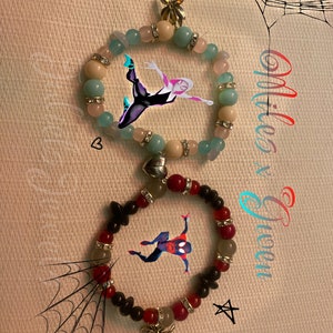 May include: Two bracelets with charms of Spider-Man characters. The top bracelet has light blue, white, and pink beads with a silver spider charm. The bottom bracelet has red, black, and white beads with a silver spider charm.