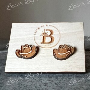 May include: A pair of wooden cowboy hat earrings on a white wooden display card with the text "Laser By B" printed on the bottom.