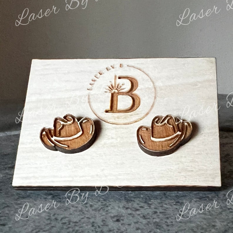 May include: A pair of wooden cowboy hat earrings on a white wooden display card with the text "Laser By B" printed on the bottom.