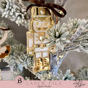 Könnte beinhalten: Goldener und weißer Nussknacker-Weihnachtsbaumschmuck, der an einem schneebedeckten künstlichen Weihnachtsbaum hängt. Der Schmuck ist aus lasergeschnittenem Acryl.