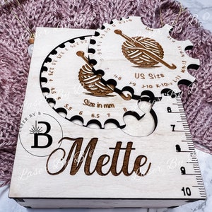 May include: A wooden box with a circular gauge for measuring knitting needle sizes. The gauge has markings for US sizes and millimeters. The box is engraved with the name "Mette" and a logo for "Laser By B".