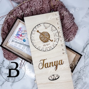 Puede incluir: Una caja de madera con una regla, un medidor de agujas de tejer y un nombre personalizado "Tanya". La caja está hecha de madera clara y tiene un acabado natural. La regla está marcada en centímetros del 1 al 25. El medidor de agujas de tejer es un gráfico circular con una bola de hilo y agujas de tejer en el centro. El medidor está marcado con tamaños de EE. UU. del 0 al 19. La caja es un gran regalo para los tejedores.