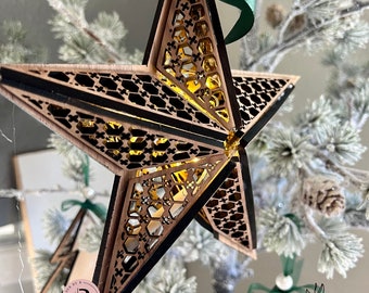 May include: A wooden star bauble with a honeycomb design and a warm glow from the lights inside. The star is hanging on a Christmas tree with green branches and white snow.