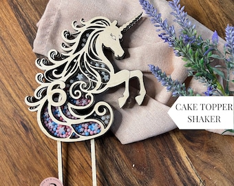 Op de afbeelding: Houten taarttopper in de vorm van een eenhoorn met een ster- en swirl-ontwerp. De manen en staart van de eenhoorn zijn uitgesneden om ruimte te creëren voor sprinkels. De topper staat op een houten stok en zegt "Cake Topper Shaker" aan de rechterkant van de afbeelding.