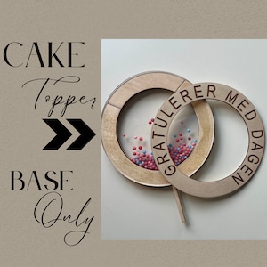 May include: Wooden cake topper with a round base and a smaller circle on top. The smaller circle has a clear plastic insert filled with pink and blue sprinkles. The larger circle has the text "GRATTULERER MED DAGEN" engraved on it.