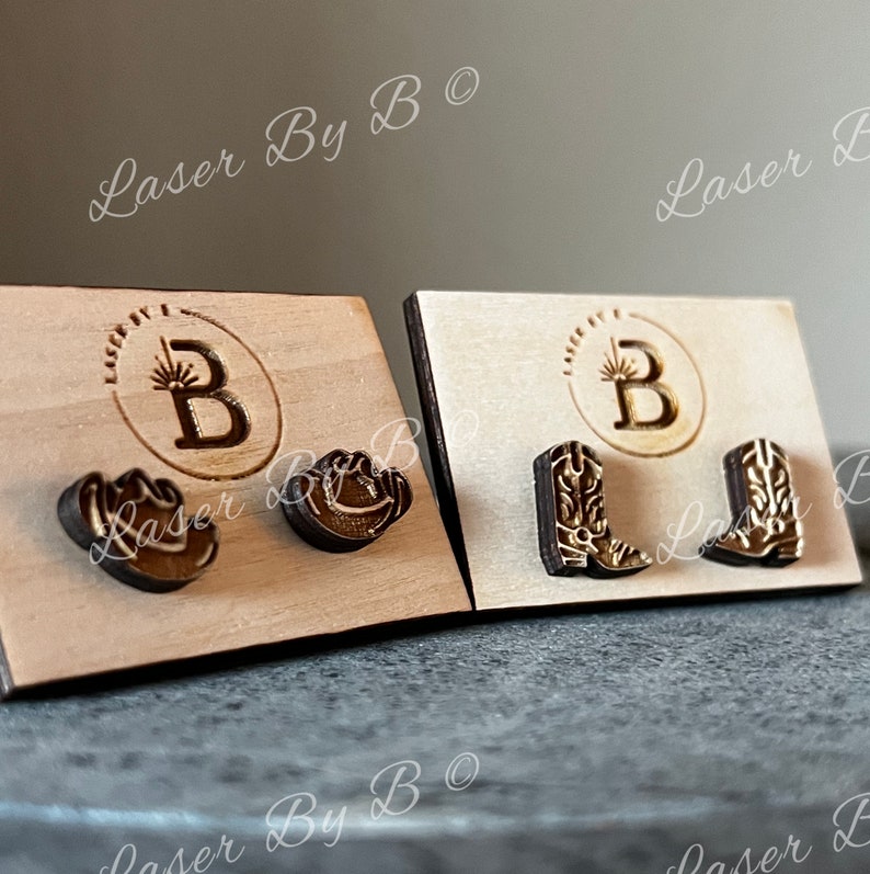 May include: Two sets of wooden earrings displayed on cards. The first set features a cowboy boot design, the second set features a cowboy hat design. The cards have the text "Laser By B" printed on them.