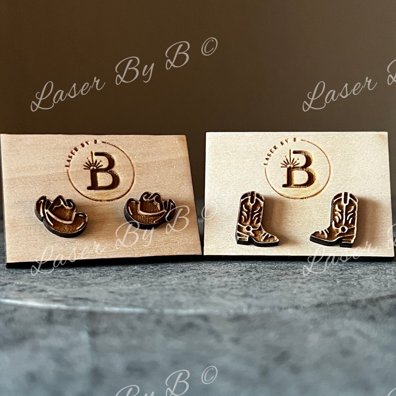 May include: Two pairs of wooden earrings displayed on a wooden card. The first pair features a cowboy hat and the second pair features a cowboy boot.