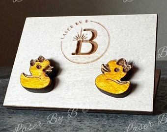 May include: A pair of wooden earrings shaped like yellow rubber duckies. The earrings are displayed on a white card with the text "Laser By B" printed on it.