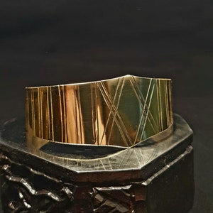 Op de afbeelding: Een gouden metalen armband met een geometrisch ontwerp. De armband is licht gebogen en heeft een gepolijste afwerking.