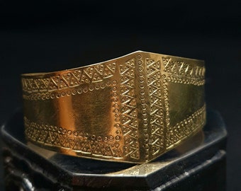 May include: A broad, gold-coloured cuff bracelet featuring geometric patterns of triangles and circles. The bracelet sits on a dark, textured surface, with a black background.