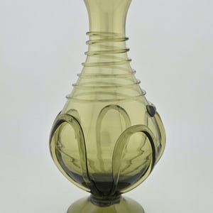 Peut inclure: Vase en verre vert olive avec un corps bulbeux et un dessus évasé. Le vase présente une série d'anneaux horizontaux autour du col et des motifs bouclés décoratifs sur les côtés. La base est un pied circulaire.