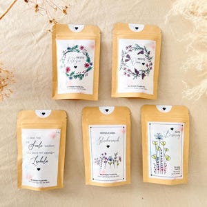 Könnte beinhalten: Fünf kleine, beigefarbene Beutel mit weißen Etiketten, die florale Designs und Text aufweisen. Die Beutel haben ein herzförmiges Detail oben. Die Etiketten zeigen Sätze wie "Beste" und "Gute Besserung" mit floralen Akzenten.