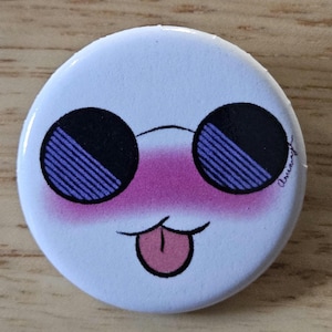 May include: A white button with a cartoon face. The face has black sunglasses with purple stripes, pink blush, and a pink tongue sticking out. The button is on a wooden surface.