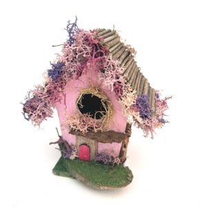 May include: A whimsical pink birdhouse with a twig roof and a small red door. The birdhouse is adorned with dried moss in shades of pink, purple, and beige. The base is covered in green moss, creating a miniature garden scene.