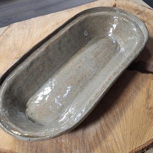 Könnte beinhalten: Eine hellgraue Keramik-Servierplatte mit gesprenkeltem Glazure. Die Platte ist oval geformt und hat einen leicht erhöhten Rand.