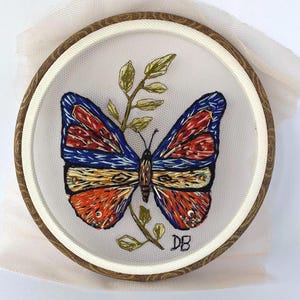 Puede incluir: Mariposa bordada con alas rojas, azules y doradas sobre un fondo blanco. La mariposa está rodeada de un tallo verde con hojas. La bordado está enmarcado en un aro de madera.