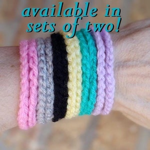 May include: A set of seven crocheted bracelets in pink, gray, black, yellow, turquoise, and lavender. The bracelets are stacked on a person's wrist. The text "available in sets of two!" is written above the bracelets.