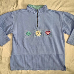 May include: A light blue quarter-zip sweatshirt with ribbed sleeves and hem. The sweatshirt features embroidered squares with a leaf, sunflower, and heart design. The collar is light green.