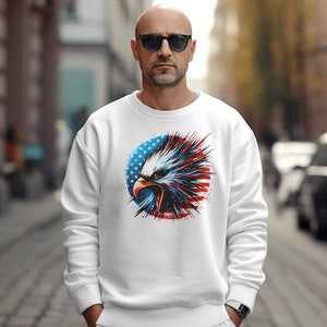 May include: White sweatshirt with a graphic of an eagle's head in front of an American flag. The eagle is red, white, and blue with a black beak and eyes.