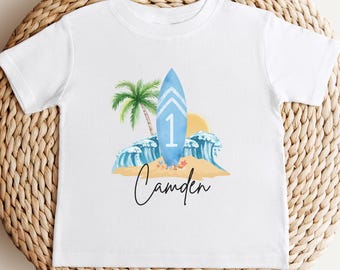 May include: White toddler t-shirt with a watercolor design featuring a blue surfboard with the number 1, a palm tree, waves, and a sun. The name "Camden" is written in script below the graphic. The shirt is displayed on a woven, circular placemat.