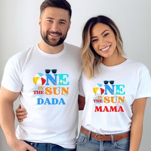 May include: Two white t-shirts with the text "ONE IN THE SUN DADA" and "ONE IN THE SUN MAMA" in blue, yellow, and red. The shirts feature a beach ball and sunglasses graphic. The people are wearing blue jeans.